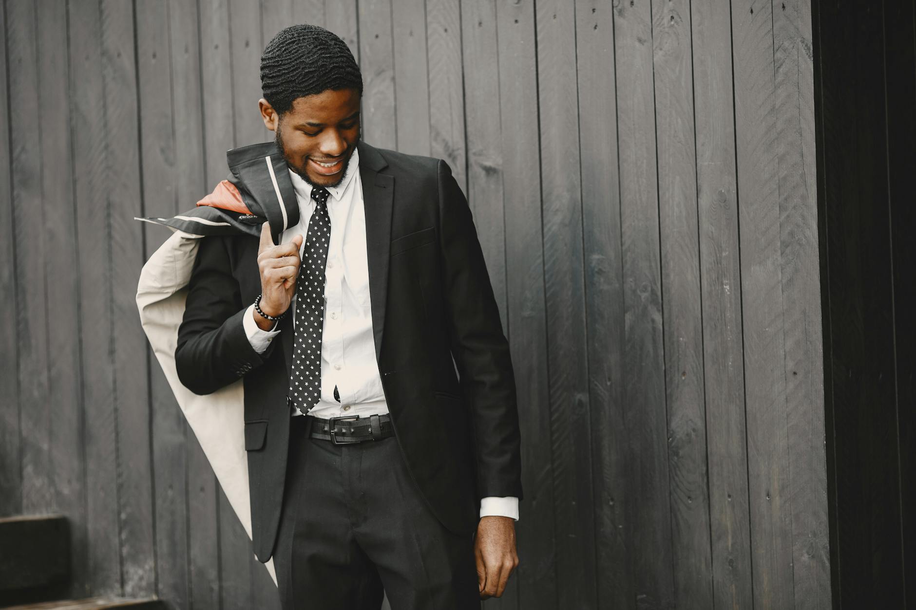 A young man in a formal suit smiling while leaning against a black wooden wall. Business portrait.